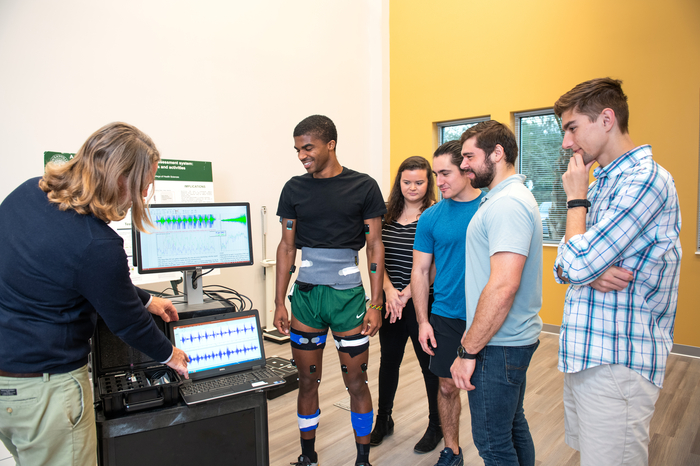 Students gathered around a machine monitoring a student's vitals listening to the professor explain the purpose of the system