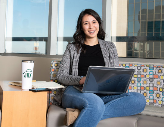 A student seating at a couch with her laptop on her hands smiles at the camera. 