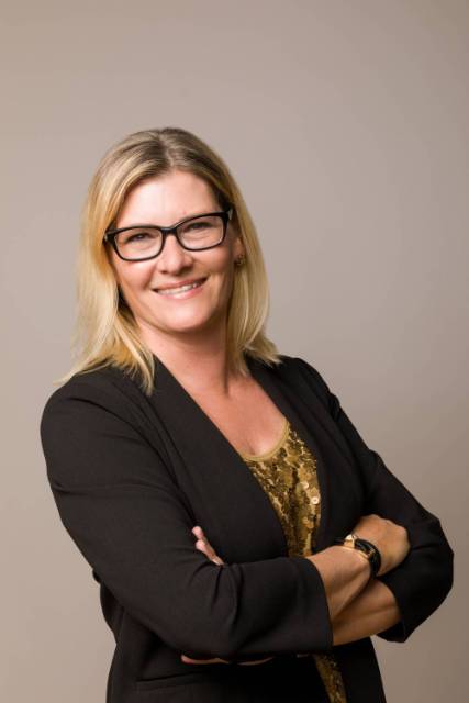 A woman with blonde and brown shoulder length hair smiling at the camera wearing black glasses, a black suit jacket, and a gold sparkly shirt