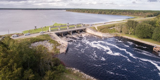 aerial view of a river dam with a grey-blue sky, cars driving over the bridge of the damn, and greenery to the sides