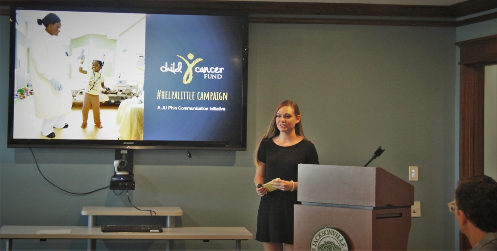 Young female stands at podium by screen with Child Cancer Fund logo