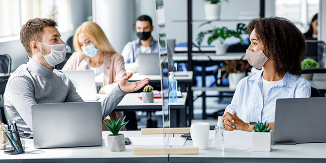 Two office employees talking through a glass partition and wearing masks.