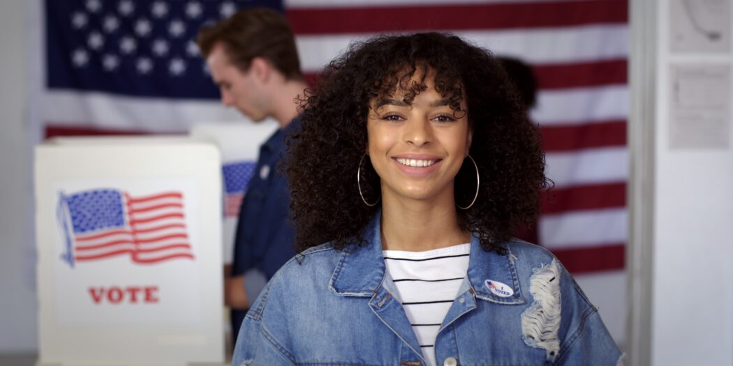 Woman wearing a jean jacket and white striped shirt standing in front of a voting booth smiling with the American flag behind her 