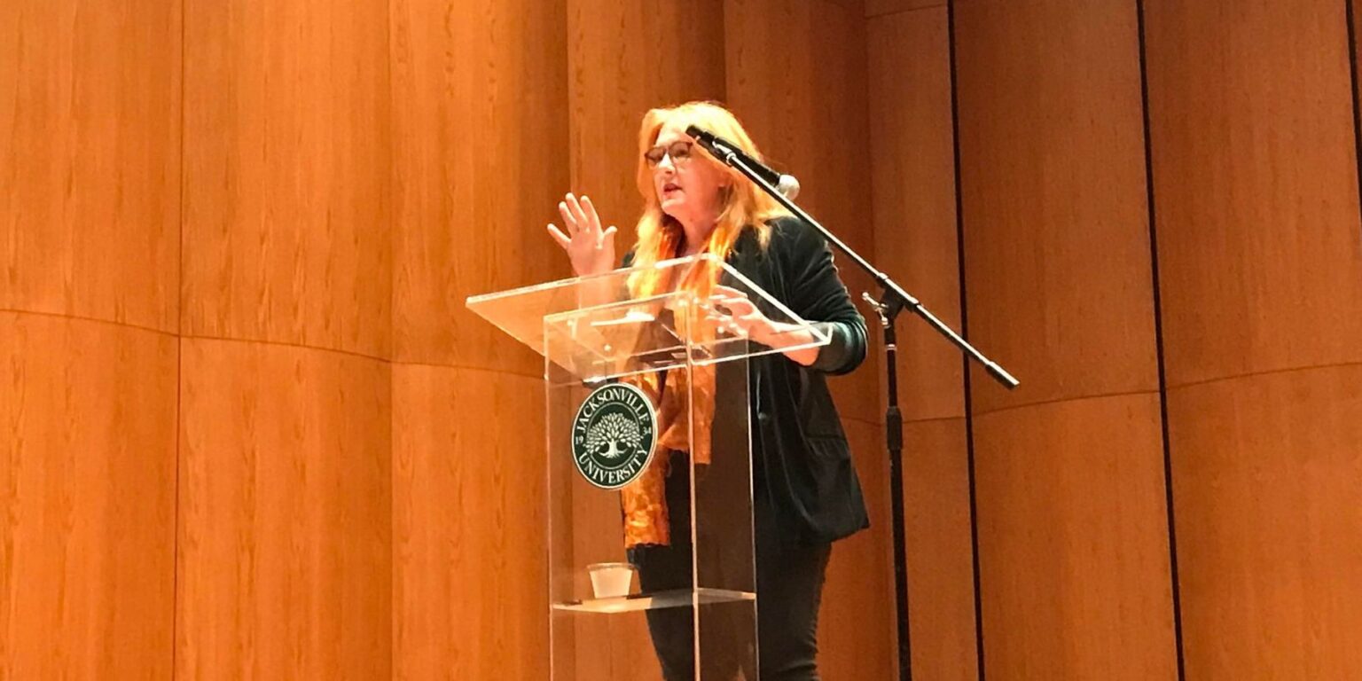 Caucasian female with blonde hair dressed in all black speaking emotively in front of a JU podium