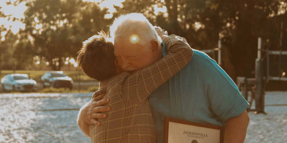 Two middle-aged adults hug at the conferring of an award against a tree-dappled, sunny, warm backdrop.