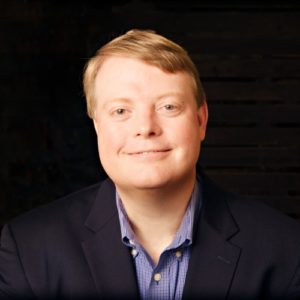 Chris hand smiles in black suit jacket and blue shirt