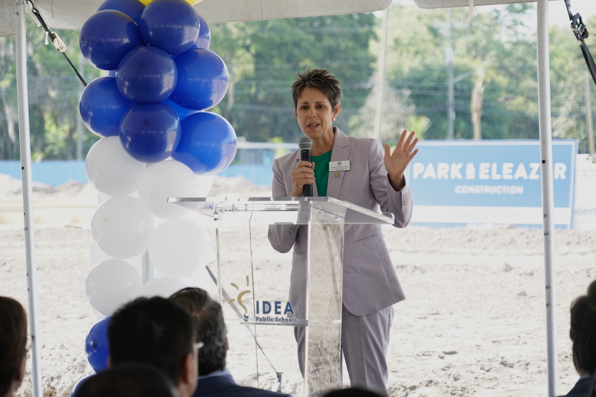 Female leader in grey suit speaking at a podium outside about the IDEA school