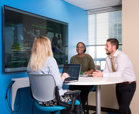 A group of people seating or standing around a table and tv monitor on the wall. 