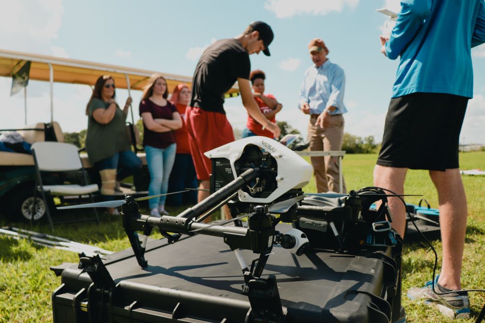 Aviation students working with a faculty member outside with drones over a green landscape