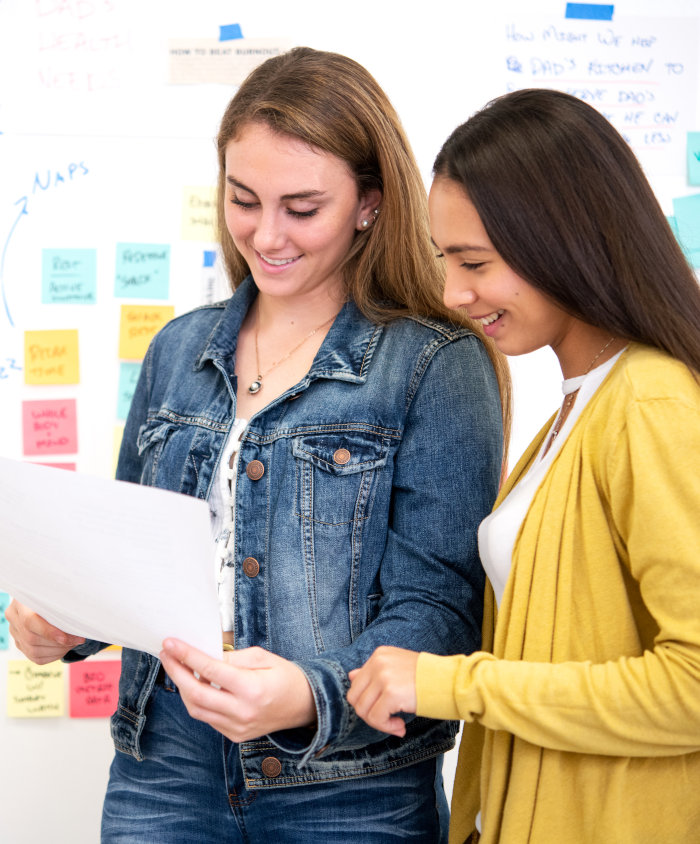 A photo of two women in front of a white board reading a document together.